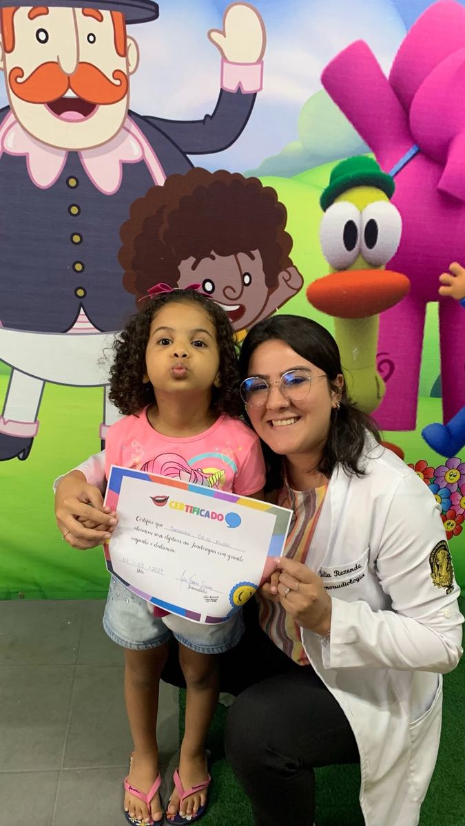 Criança recebendo certificado de conclusão da terapia ao lado da fonoaudióloga Júlia